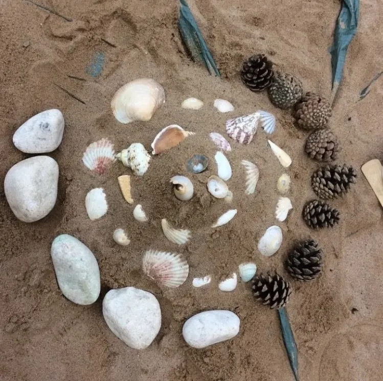 Image shows a spiral of shells and stones on sand at the beach.