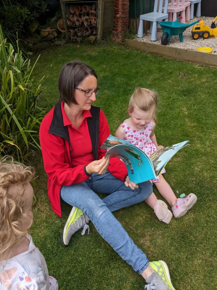 Woman sat on grass reading book with two early years children
