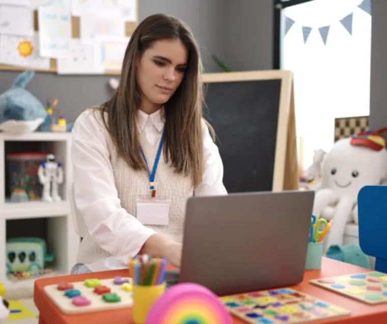 An early years practitioner sat in a nursery classroom typing on a laptop