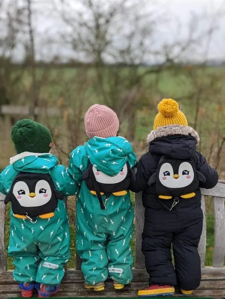 Photo of the back of three children wearing puddle suits and penguin backpacks, standing on a bench looking out over a field