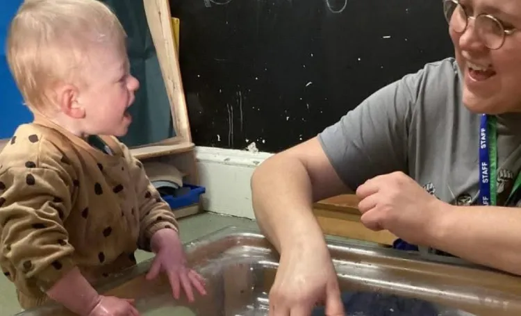 An Early Years Practitioner communicating with a child over a play table in a nursery setting. 