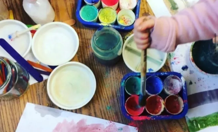 Pots of paint and paper on a table with a child's hand holding a paintbrush.
